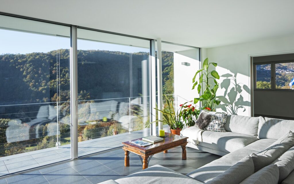 Wohnzimmer mit grauer Couch und großer Fensterfront aus Glas, mit Blick auf Wälder.